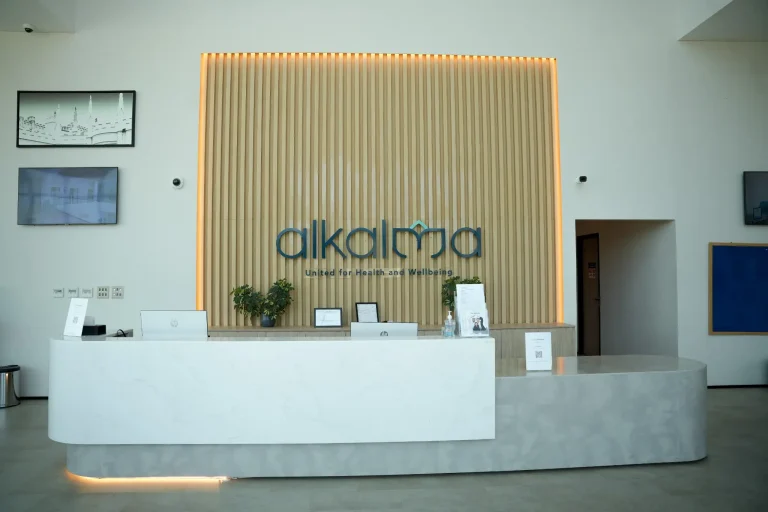 Reception desk at the alkalma Al Bateen clinic beneath a backlit alkalma logo on a slatted timber wall